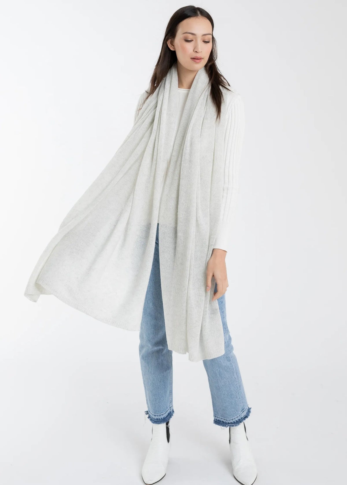 A woman modeling a long, lightweight off-white knit scarf draped over her shoulders, paired with light blue jeans and white ankle boots against a plain white studio background.