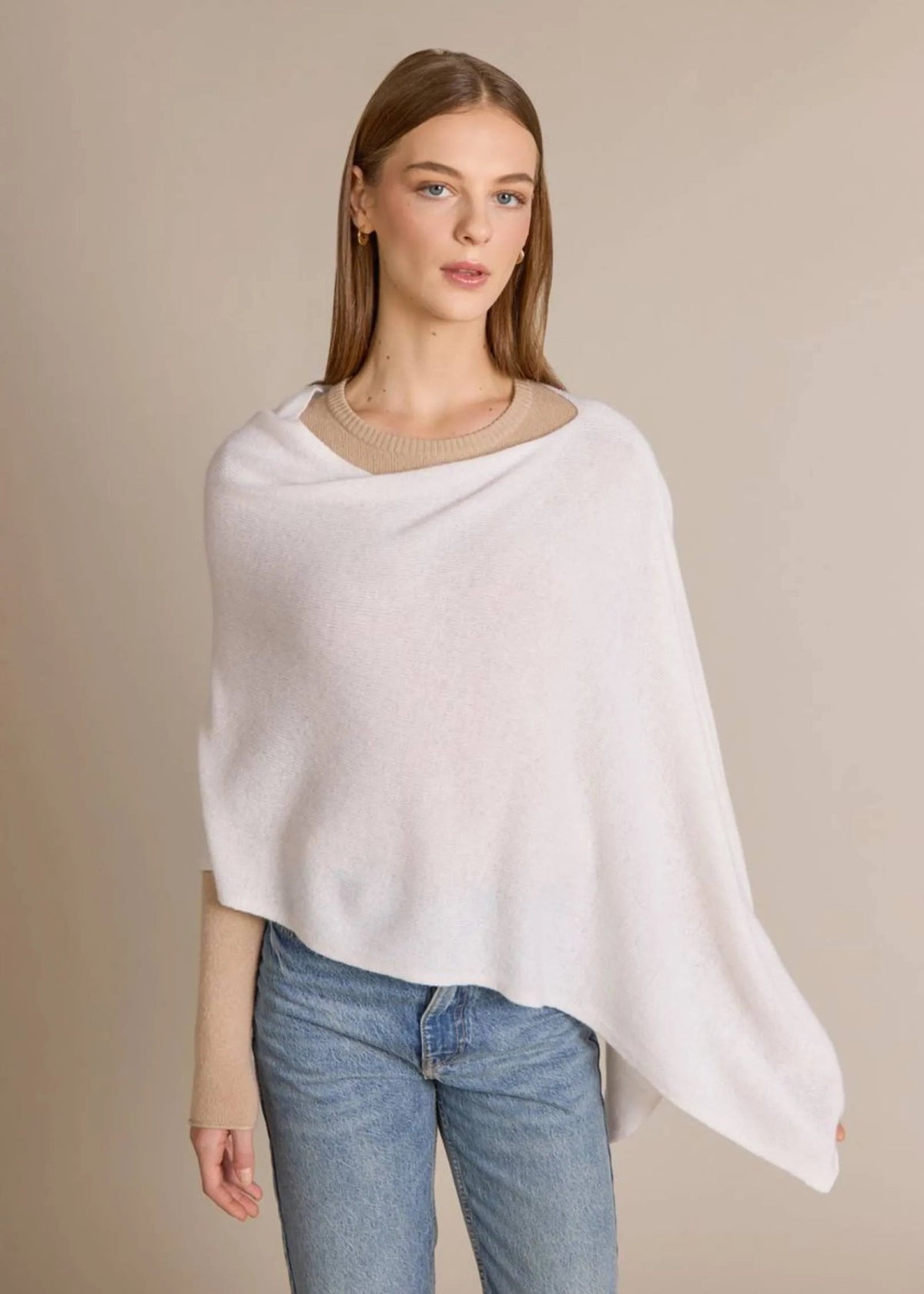 A woman modeling a light cream knit poncho over a beige crewneck sweater and blue jeans, set against a solid beige studio background.