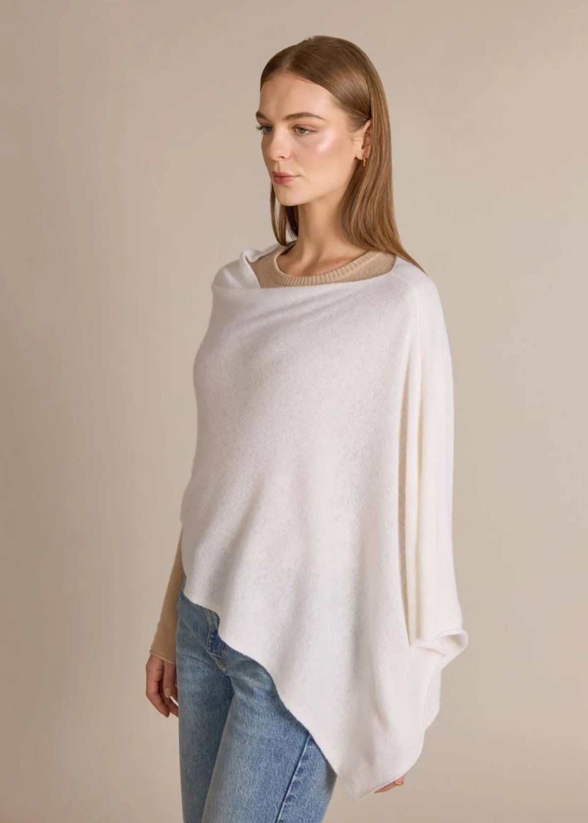 A woman wearing a soft, off-white knit poncho sweater over a beige long-sleeve top and blue jeans, standing against a solid beige studio background.