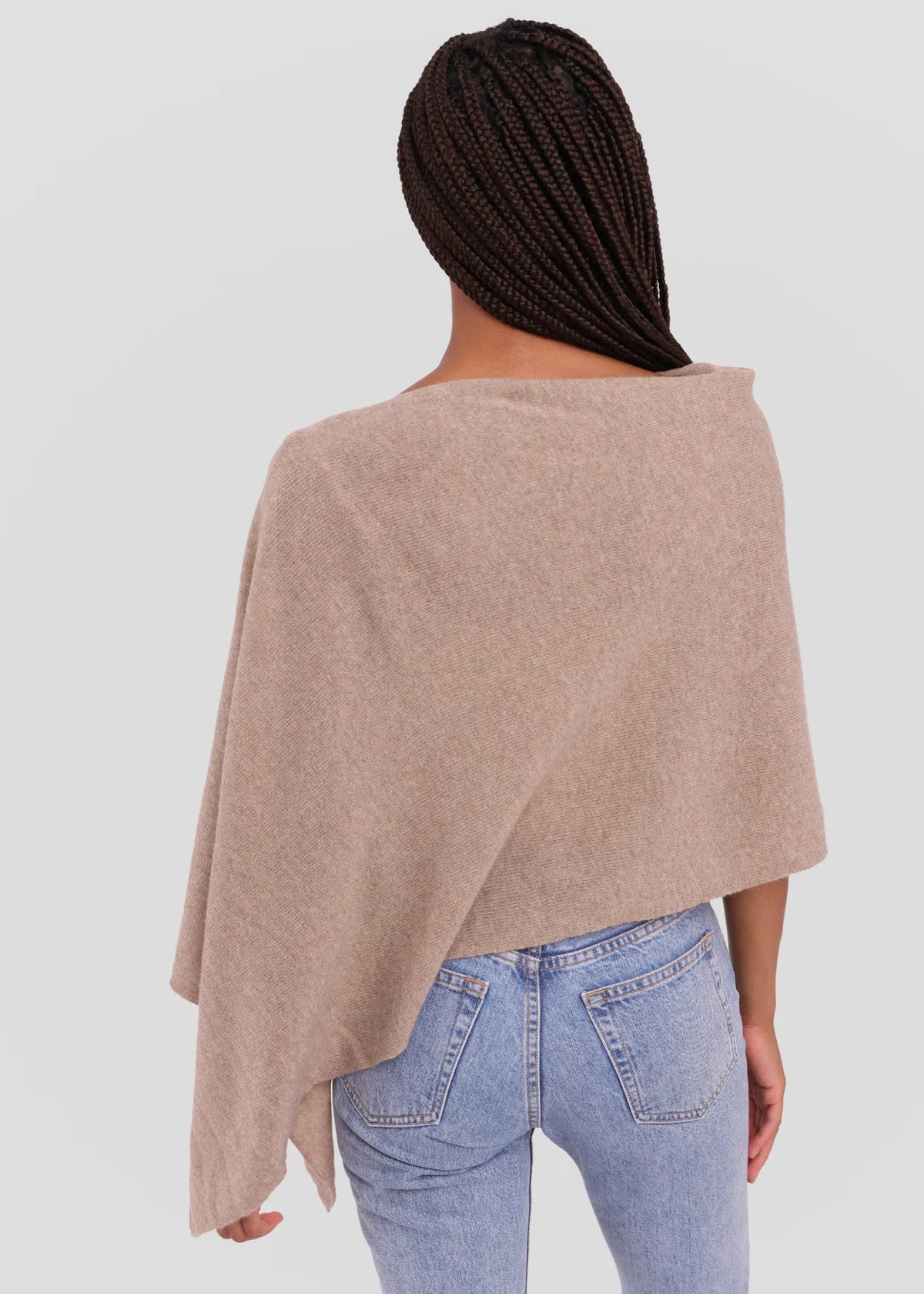 A woman viewed from behind wearing a light taupe knit poncho over light blue jeans, set against a solid light gray background.
