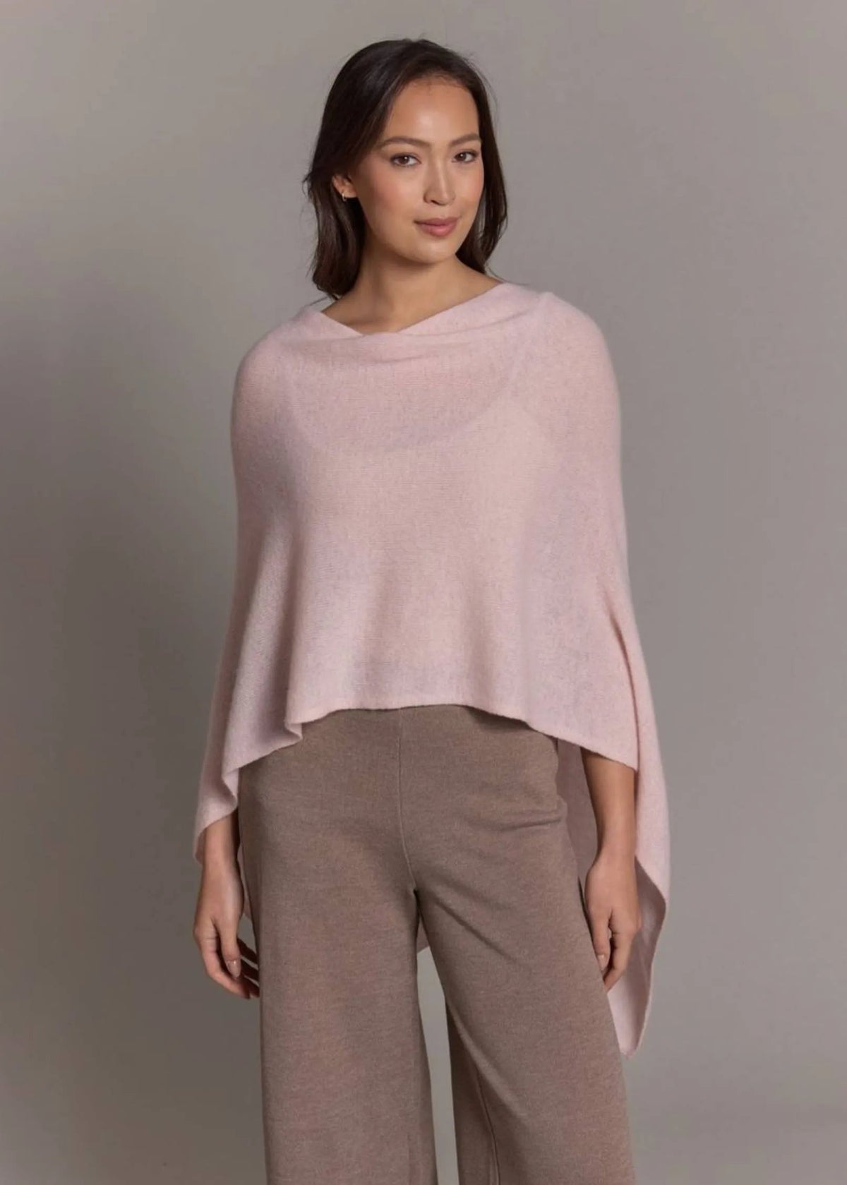 Woman wearing a light pink poncho over a beige outfit against a gray background
