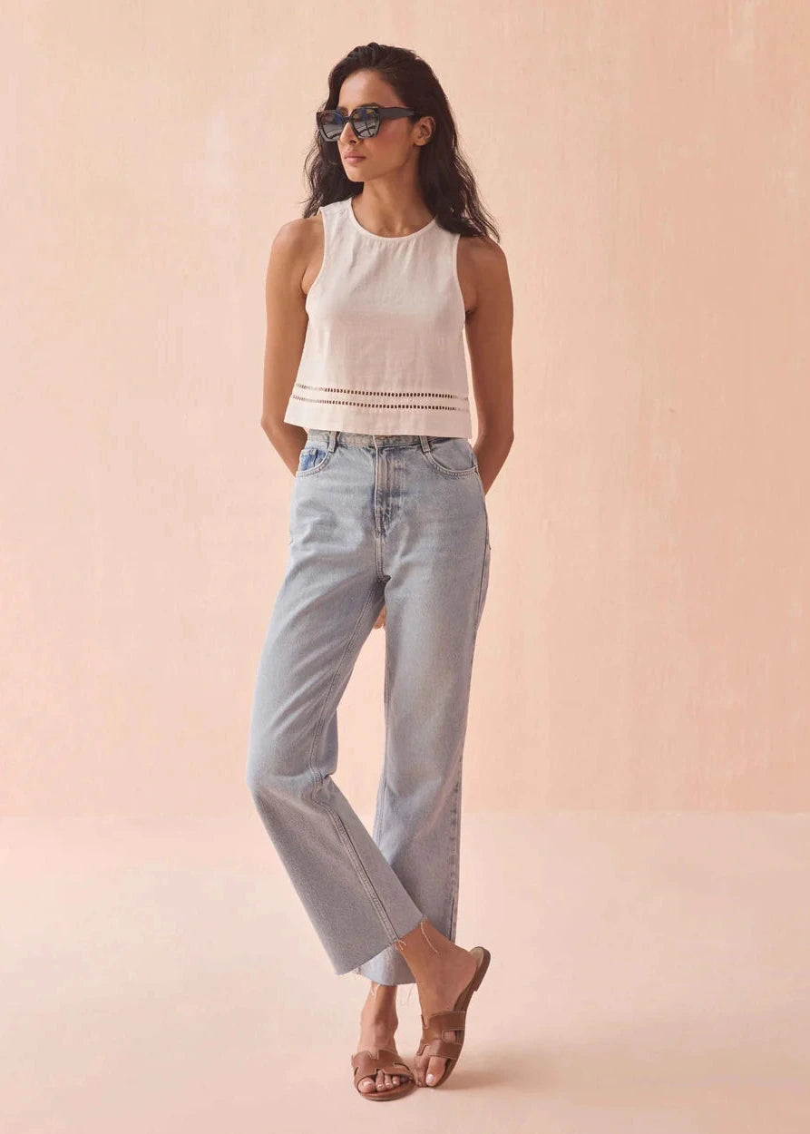 Woman wearing a white sleeveless top and light blue jeans against a pink background