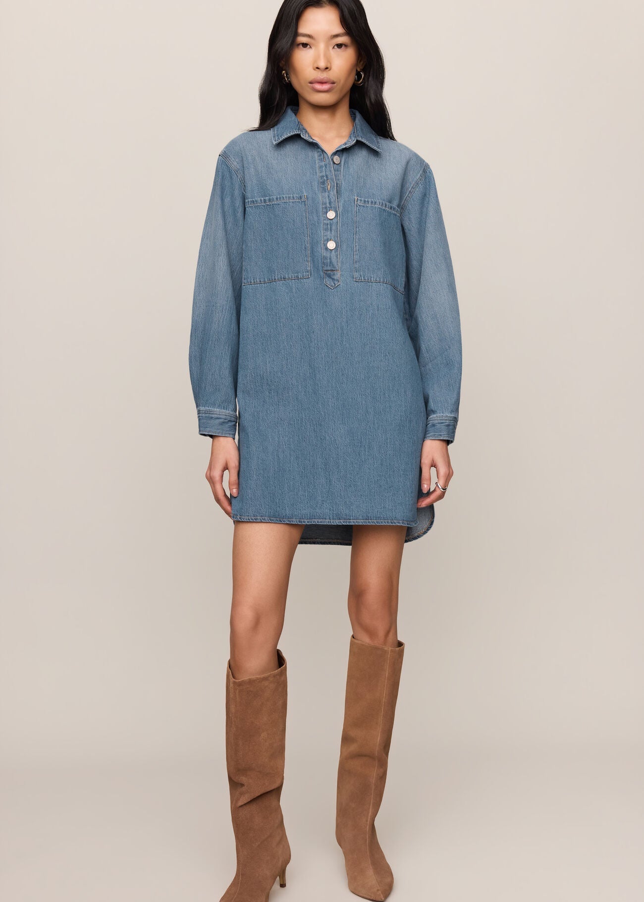 A model wearing a medium wash blue denim shirt dress with brown suede knee-high boots, standing against a solid beige studio background.