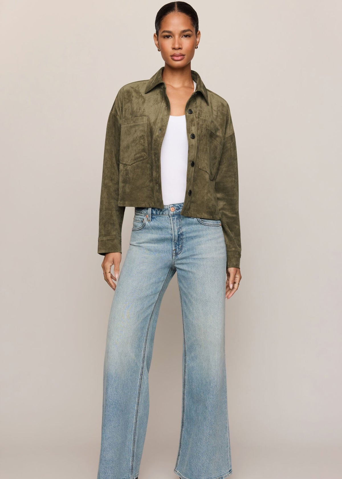 A model stands against a plain beige studio background wearing an olive green cropped suede jacket over a white top, paired with light-wash wide-leg blue jeans and black boots.