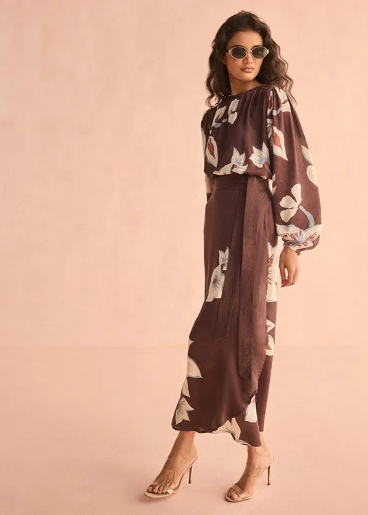 A woman modeling a brown long-sleeve maxi dress with a white floral print, styled with gold high-heeled sandals and sunglasses against a solid pink background.