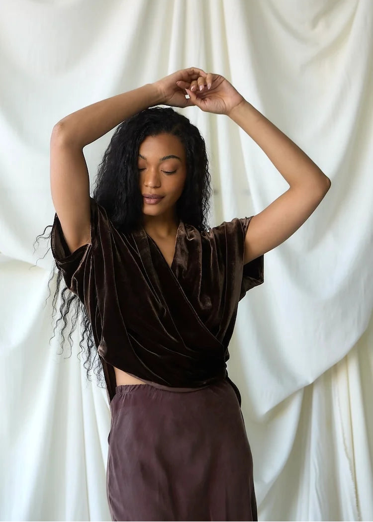 A woman with long, curly black hair poses in a dark brown velvet short-sleeve wrap top and a matching dark brown skirt against a draped white fabric backdrop.