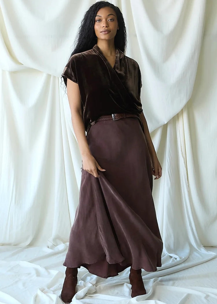 A woman stands in front of draped white fabric, wearing a dark brown velvet short-sleeve top and a matching dark brown satin maxi skirt with a thin brown belt.