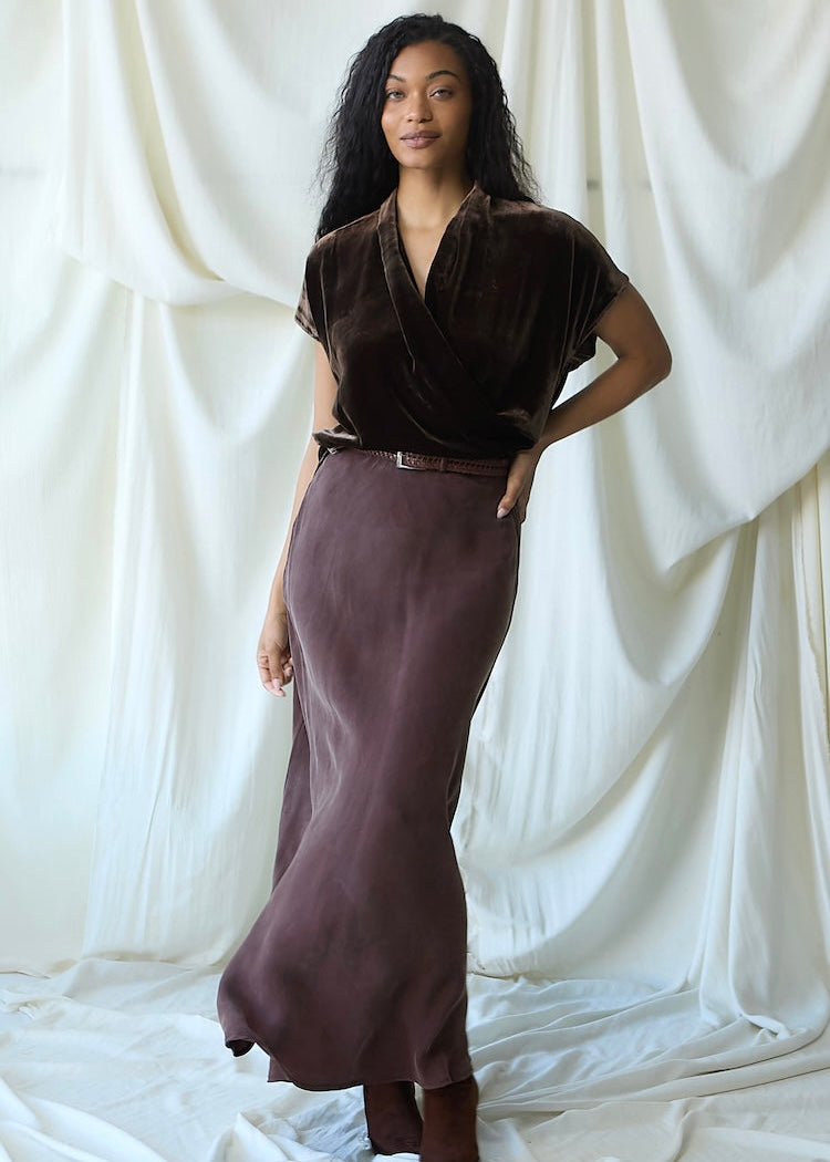 A woman modeling a dark brown velvet short-sleeve wrap top and a matching dusty mauve-brown satin midi skirt, styled with a thin brown belt and brown boots, set against a draped white fabric backdrop.