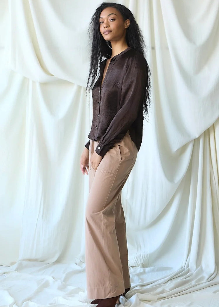A woman stands in a studio wearing a dark brown button-up shirt and beige wide-leg pants, posed against a draped white fabric backdrop.
