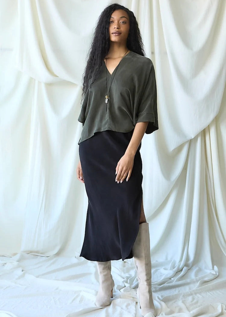 A woman stands against a draped white fabric backdrop wearing an olive green short-sleeve top with a front zipper, a dark navy midi skirt, and light beige knee-high boots.