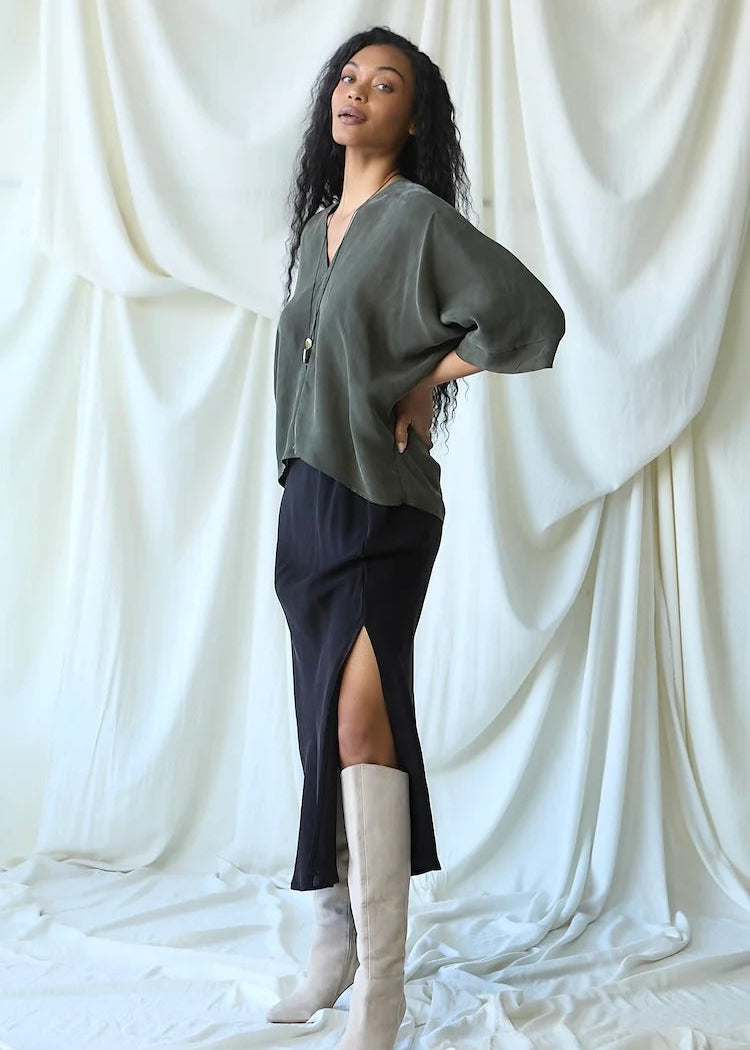A woman stands in front of draped white fabric, wearing a loose olive green top with wide sleeves, a dark navy slit skirt, and tall off-white knee-high boots.