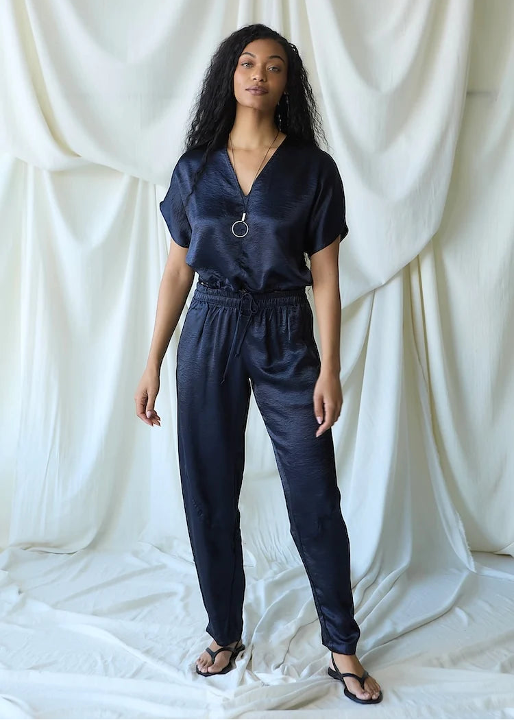 A woman modeling a dark navy blue satin-like jumpsuit with a V-neck, short sleeves, and a drawstring waist, standing against a draped white fabric backdrop.