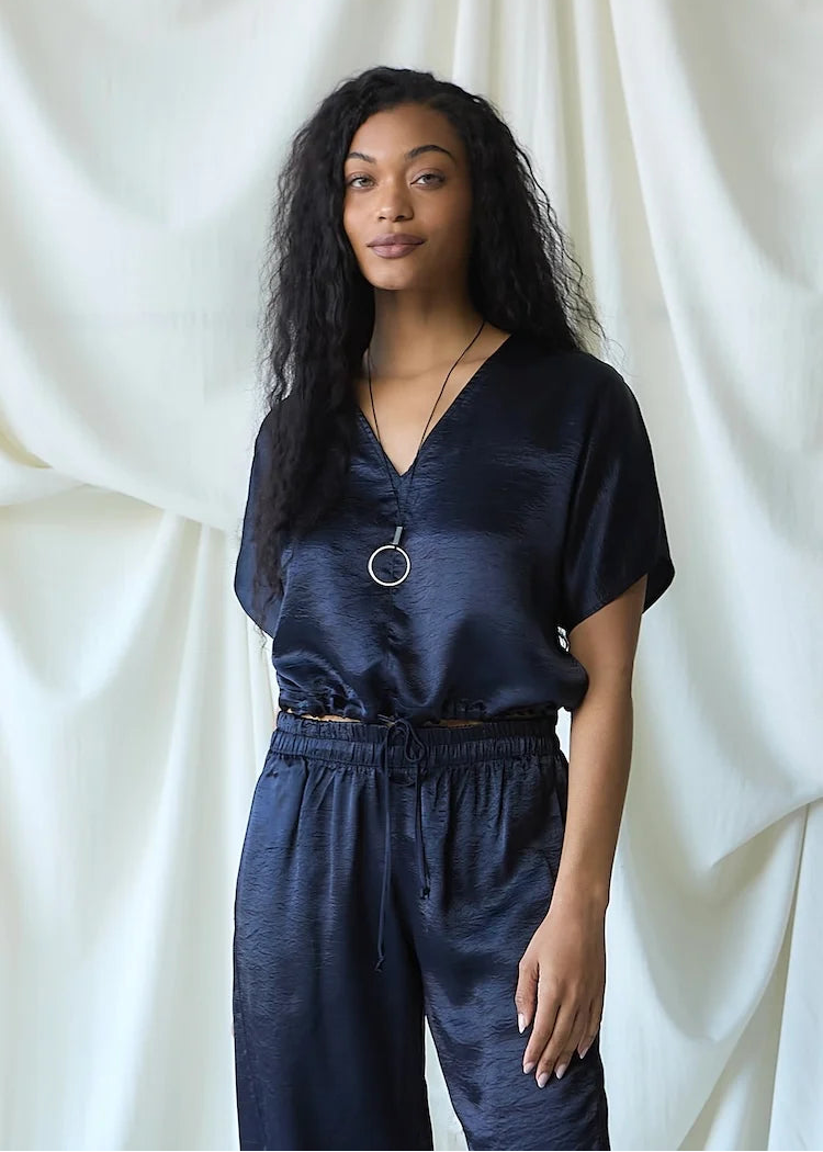 A woman with long, curly black hair stands against a draped white fabric backdrop, wearing a dark navy blue satin-like short-sleeve top and matching drawstring pants.