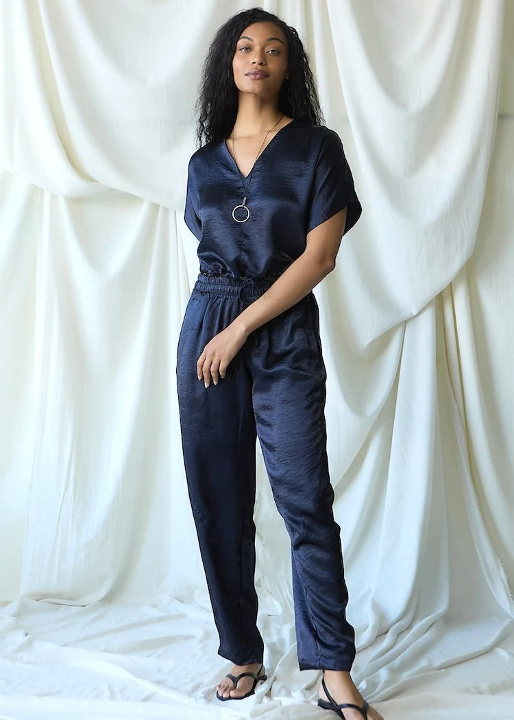 A woman modeling a dark navy blue satin jumpsuit with a V-neckline, short sleeves, and an elastic waist, standing against a draped white fabric backdrop.