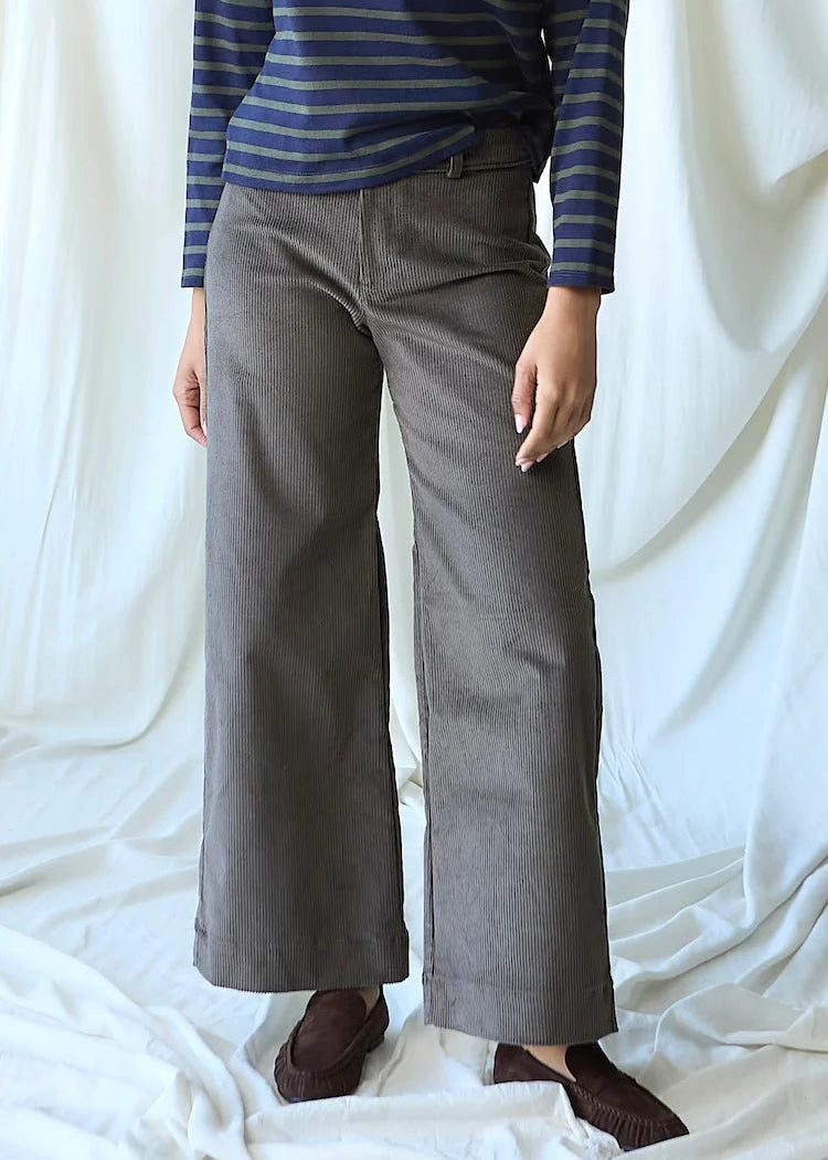 A person wearing wide-leg gray trousers, a blue and navy striped long-sleeve shirt, and brown slip-on shoes, standing against a draped white fabric backdrop.