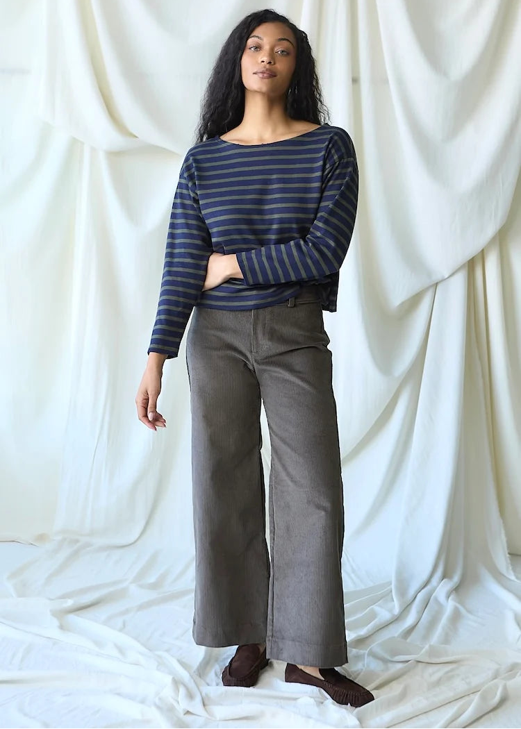A woman stands against a draped white fabric backdrop wearing a navy and blue striped long-sleeve top with dark gray wide-leg pants and brown shoes.