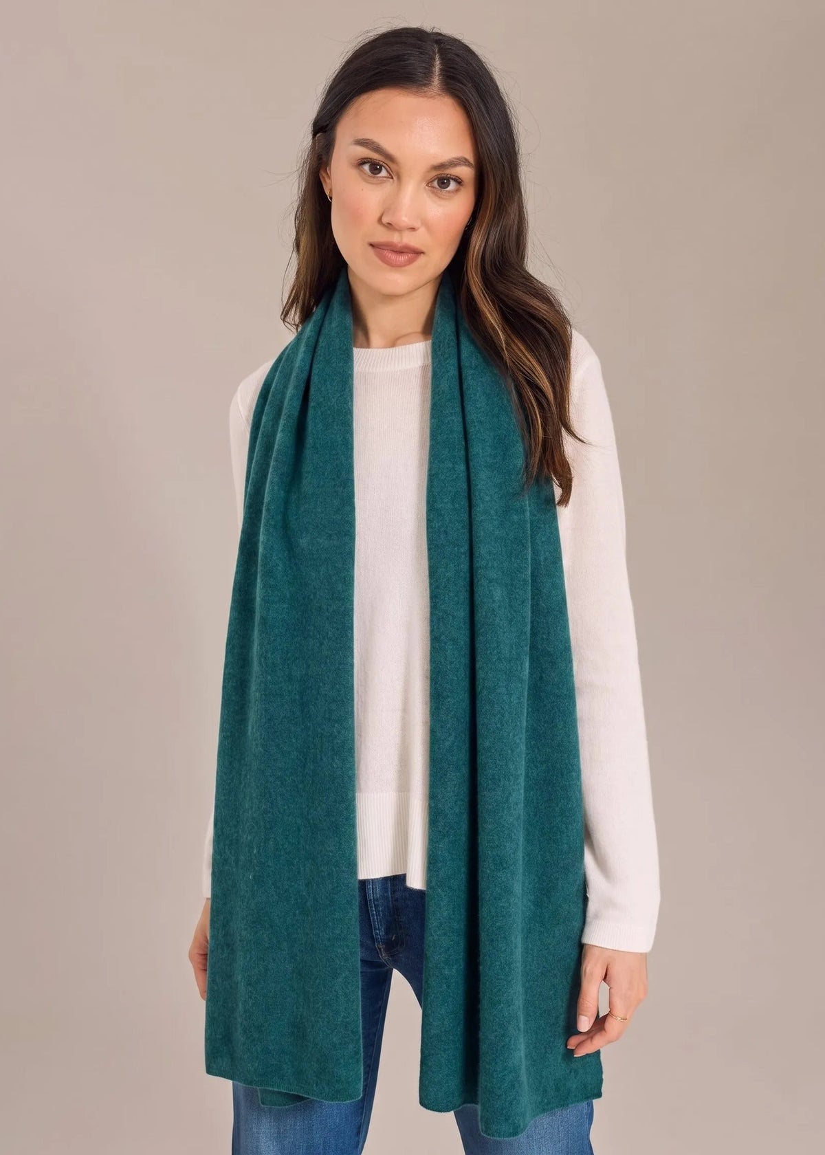 A woman modeling a long teal green scarf draped over her shoulders, paired with a white long-sleeve top and blue jeans, against a solid beige studio background.