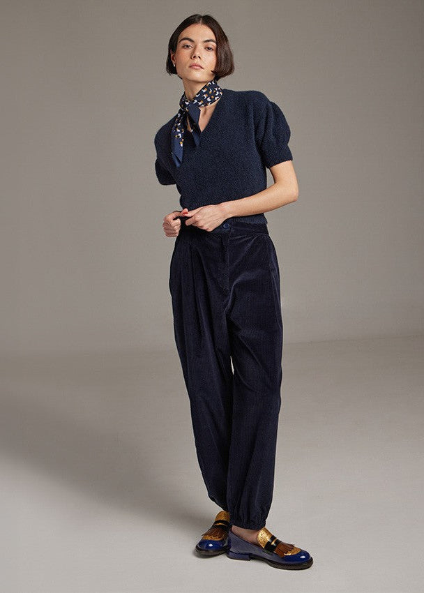 A model stands in a studio wearing a dark navy blue knit short-sleeve top with a split neckline, matching navy corduroy trousers, a patterned navy neck scarf, and dark loafers with gold and blue accents.