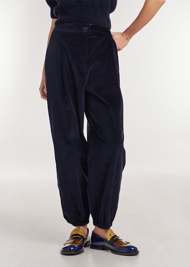 A person wearing dark navy corduroy trousers with a relaxed, baggy fit and elastic cuffs, paired with a dark top and blue leather loafers with gold detailing, against a plain light gray studio background.