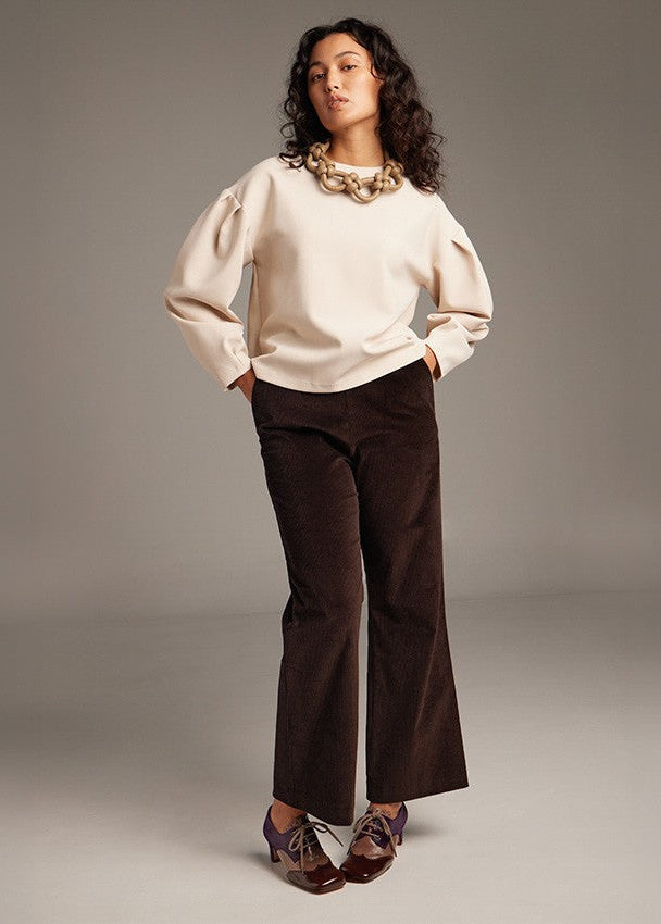 A model stands against a neutral gray studio background wearing a cream puff-sleeve top, dark brown wide-leg corduroy pants, and brown lace-up shoes with a chunky gold necklace.
