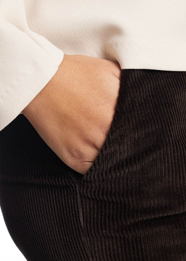 Close-up of a person wearing dark brown corduroy pants with a white top, with the hand in the pocket.
