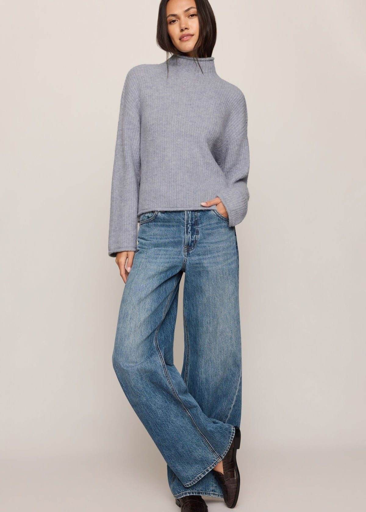 A model wearing a light gray ribbed mock-neck sweater and wide-leg blue jeans, standing against a plain beige studio background.