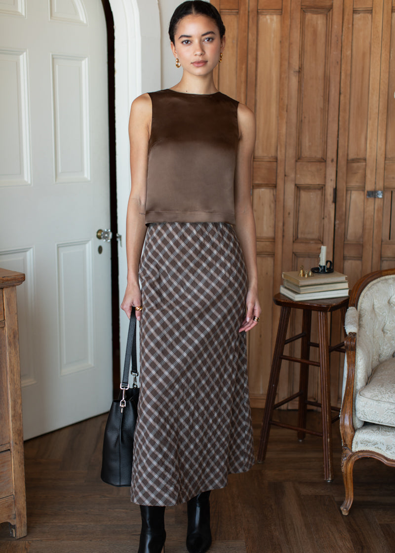 Woman in a brown sleeveless top and checkered skirt standing in a room with wooden furniture.