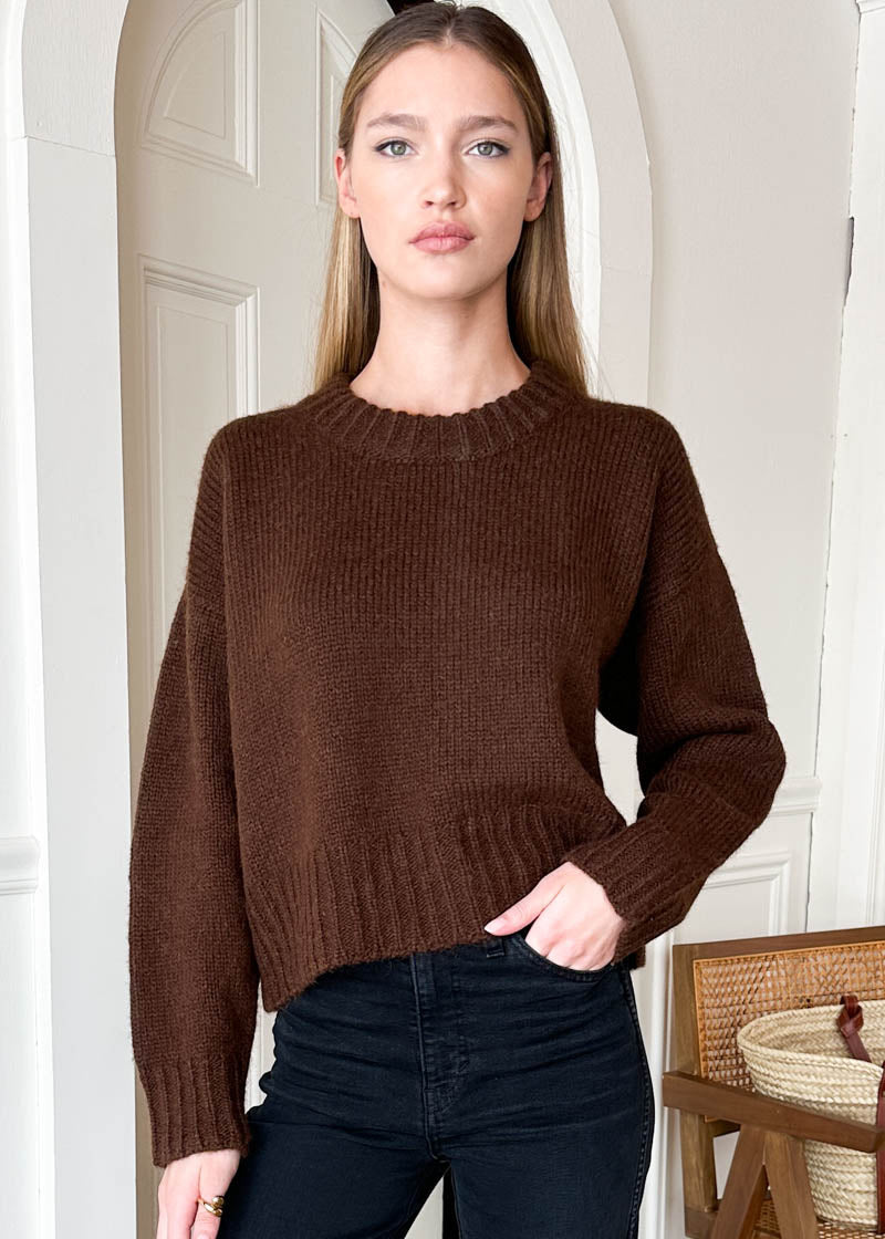 A woman wearing a brown ribbed knit sweater and black jeans, standing in front of a white paneled door with a wooden chair partially visible on the right.