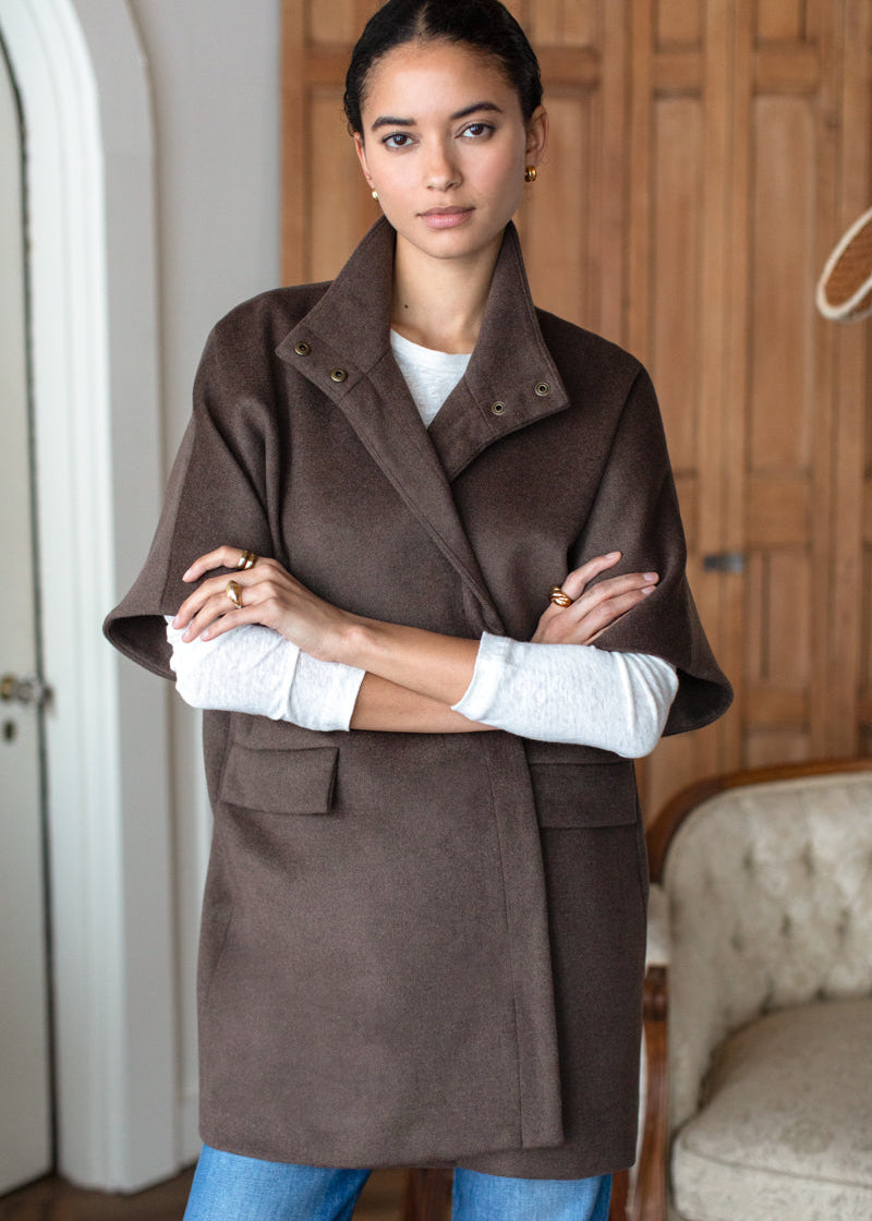 A woman wearing a brown wool cape-style coat over a white long-sleeve top and blue jeans, standing in a room with wooden doors and a tufted chair.