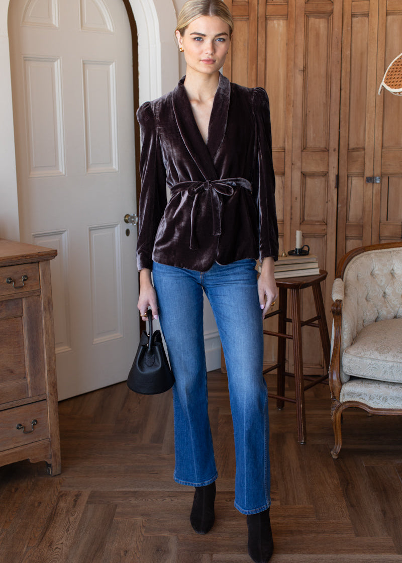Woman wearing a dark velvet top and blue jeans in a room with wooden floors and furniture.