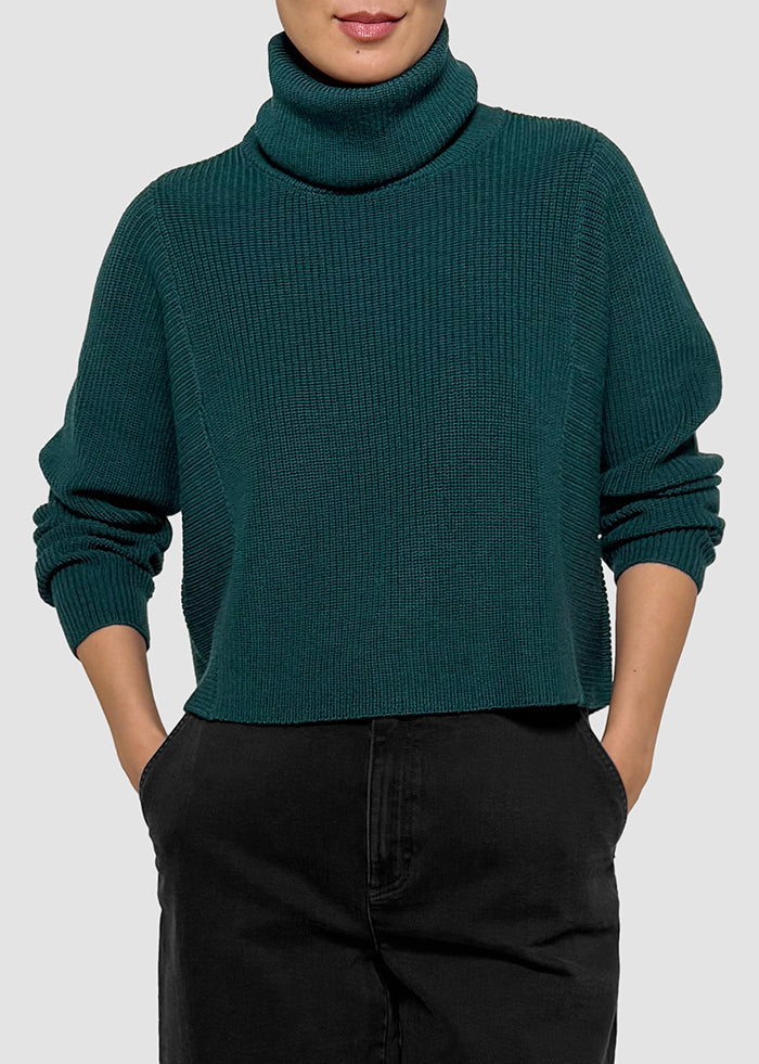 A person wearing a dark teal ribbed knit turtleneck sweater with black pants, shown from the mouth down against a light gray studio background.