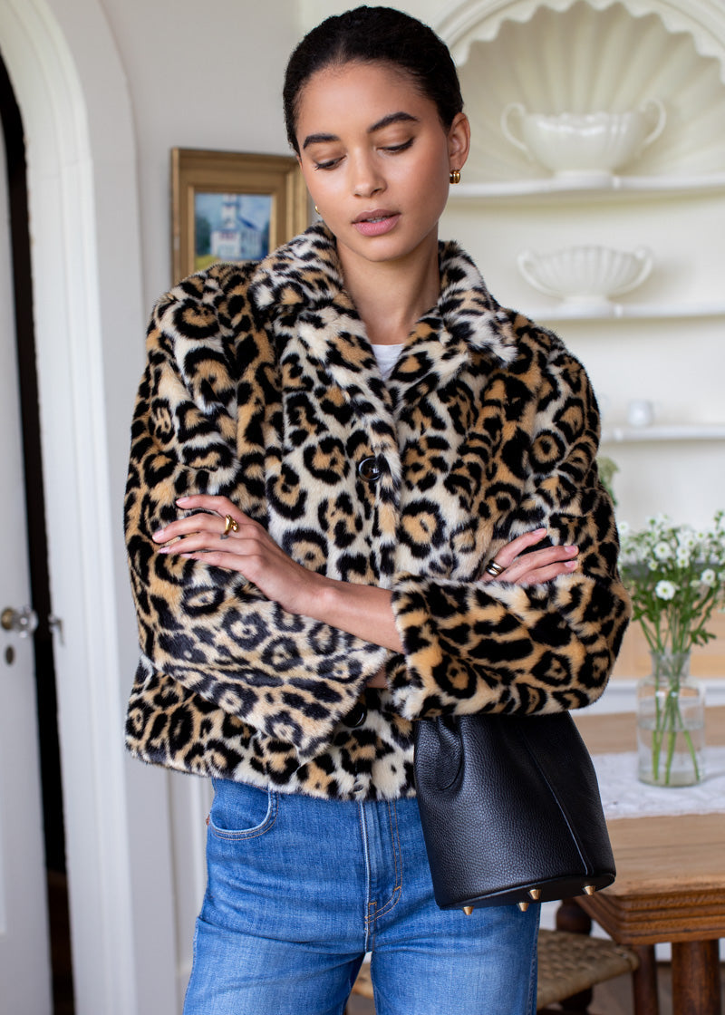 A woman wearing a plush faux-fur jacket with a leopard print pattern in tan, brown, and black, paired with blue jeans and a black handbag, standing in a bright indoor setting.