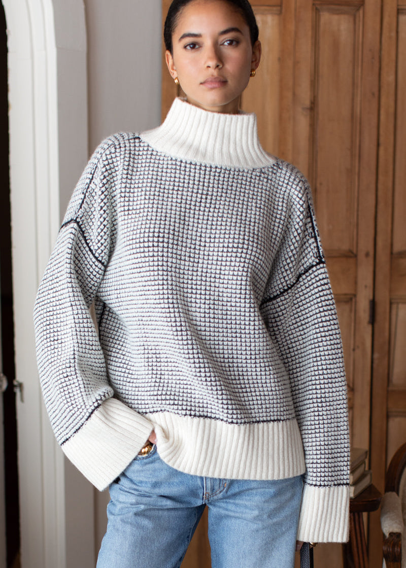Woman wearing a black and white patterned sweater with a white turtleneck underneath, standing indoors.