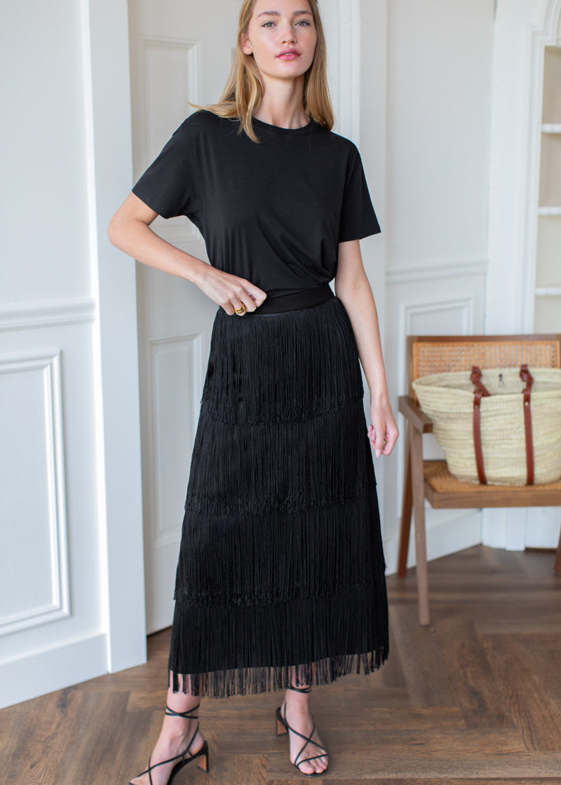 A woman modeling a black short-sleeve midi dress with a tiered fringe skirt, standing in a bright room with white walls and a wooden floor.