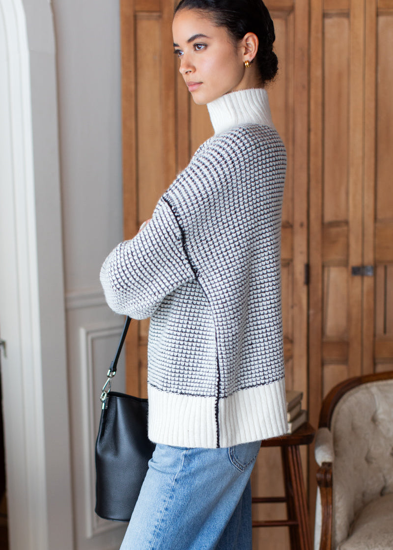 A woman wearing a black and white textured knit turtleneck sweater, paired with light blue jeans and carrying a black leather shoulder bag, standing in front of a wooden door.