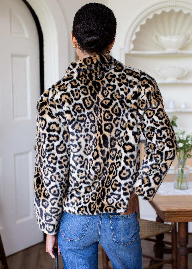 A person shown from behind wearing a plush faux-fur cropped jacket with a leopard print pattern in beige, brown, and black, paired with blue jeans in an indoor home setting.