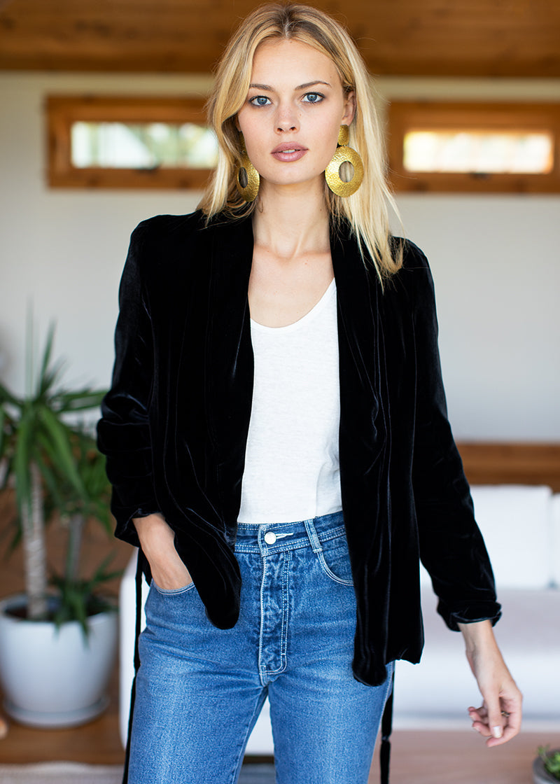 A woman wearing a black velvet blazer over a white tank top and blue jeans, standing in a brightly lit indoor setting with a blurred background.