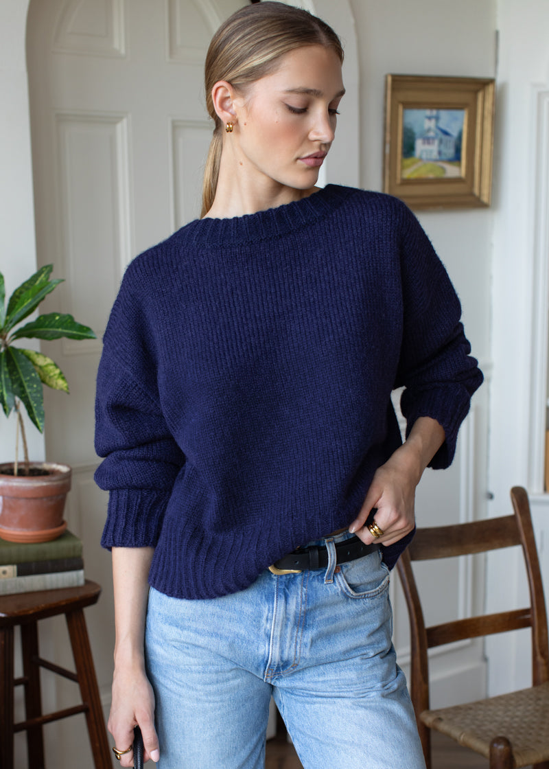 A woman wearing a navy blue ribbed knit sweater and light wash blue jeans, standing in a home interior setting.