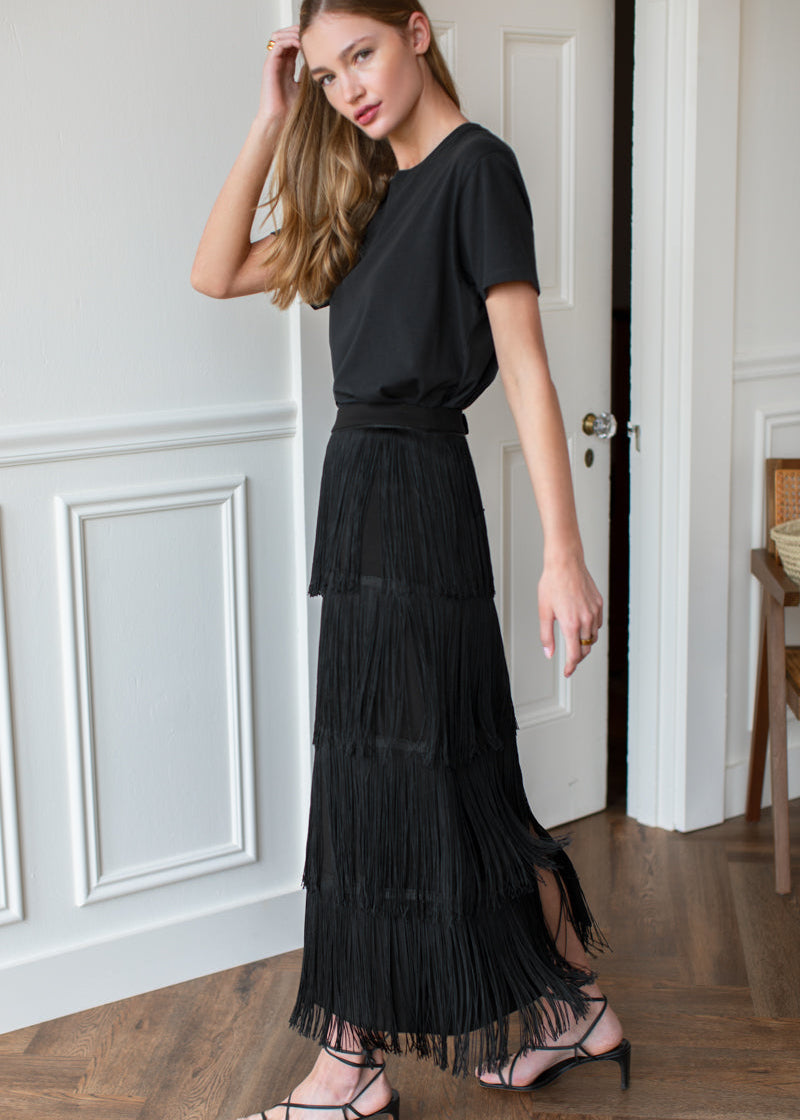 A woman modeling a black short-sleeve top and a long, tiered fringe maxi skirt, paired with strappy high-heeled sandals, standing in a room with white paneled walls and a wooden floor.