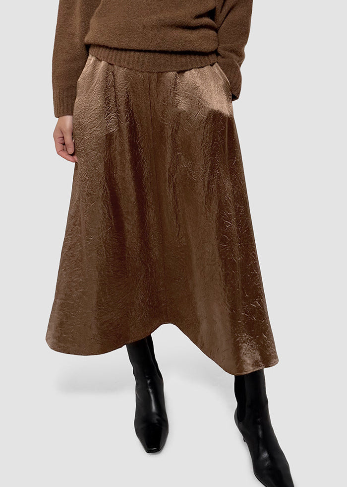 A model wearing a brown textured midi skirt with a gathered waist, paired with a brown sweater and black ankle boots, shown from the chest down on a light gray background.