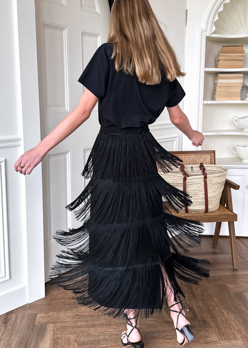 A woman wearing a black short-sleeve top and a long, tiered black pleated maxi skirt with fringe, shown from the back in a bright room with a wooden chair and woven bag nearby.