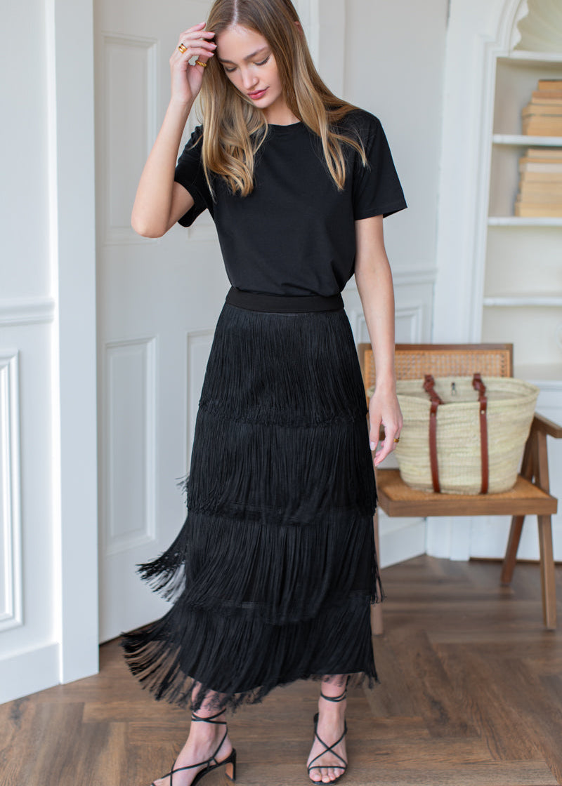 A woman models a black outfit consisting of a short-sleeve crewneck t-shirt and a long, tiered fringe midi skirt, paired with strappy heels, in a bright room with a wooden floor and a woven bag on a chair.