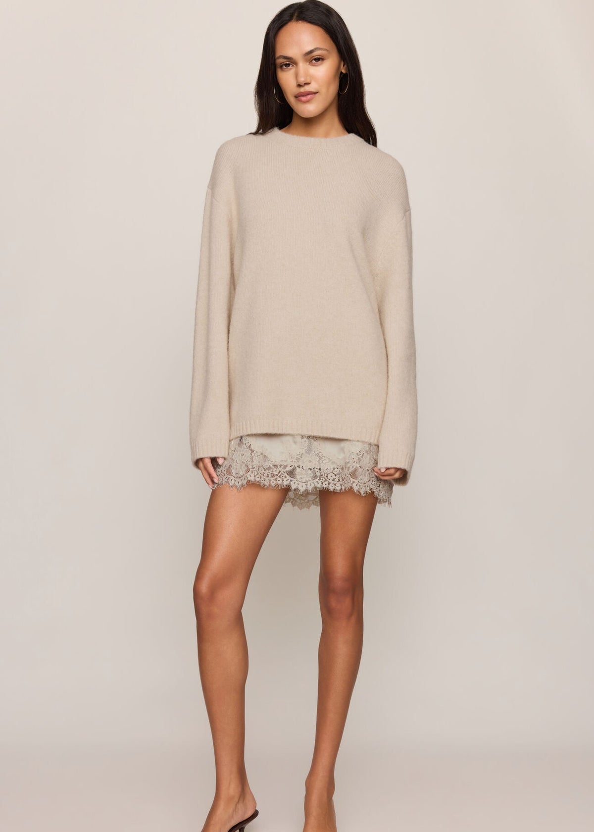 A woman modeling a light beige knit sweater dress with a short lace hem, paired with clear strap high-heeled sandals, against a solid light beige studio background.