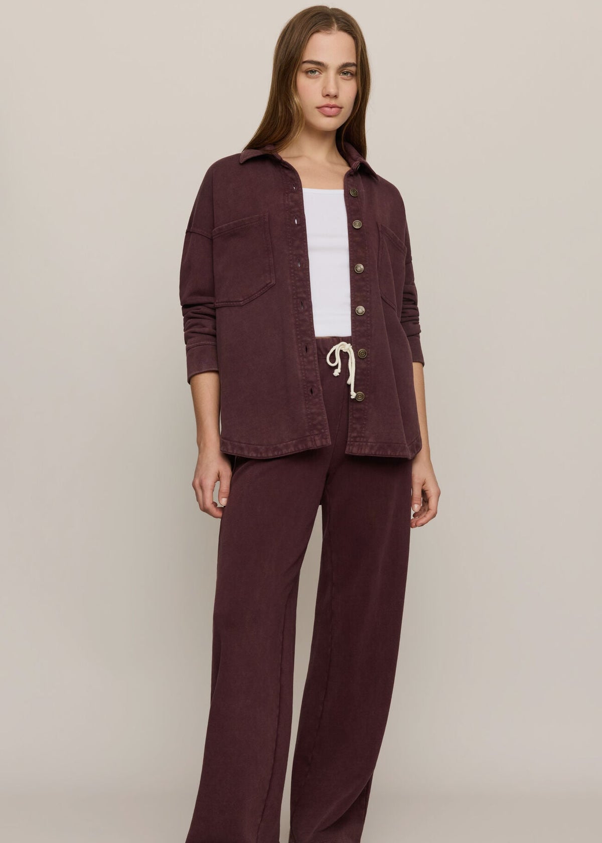 A woman modeling a matching two-piece lounge set in a deep burgundy color, consisting of a button-up shirt-jacket and wide-leg pants, styled over a white top and brown slippers against a neutral studio background.