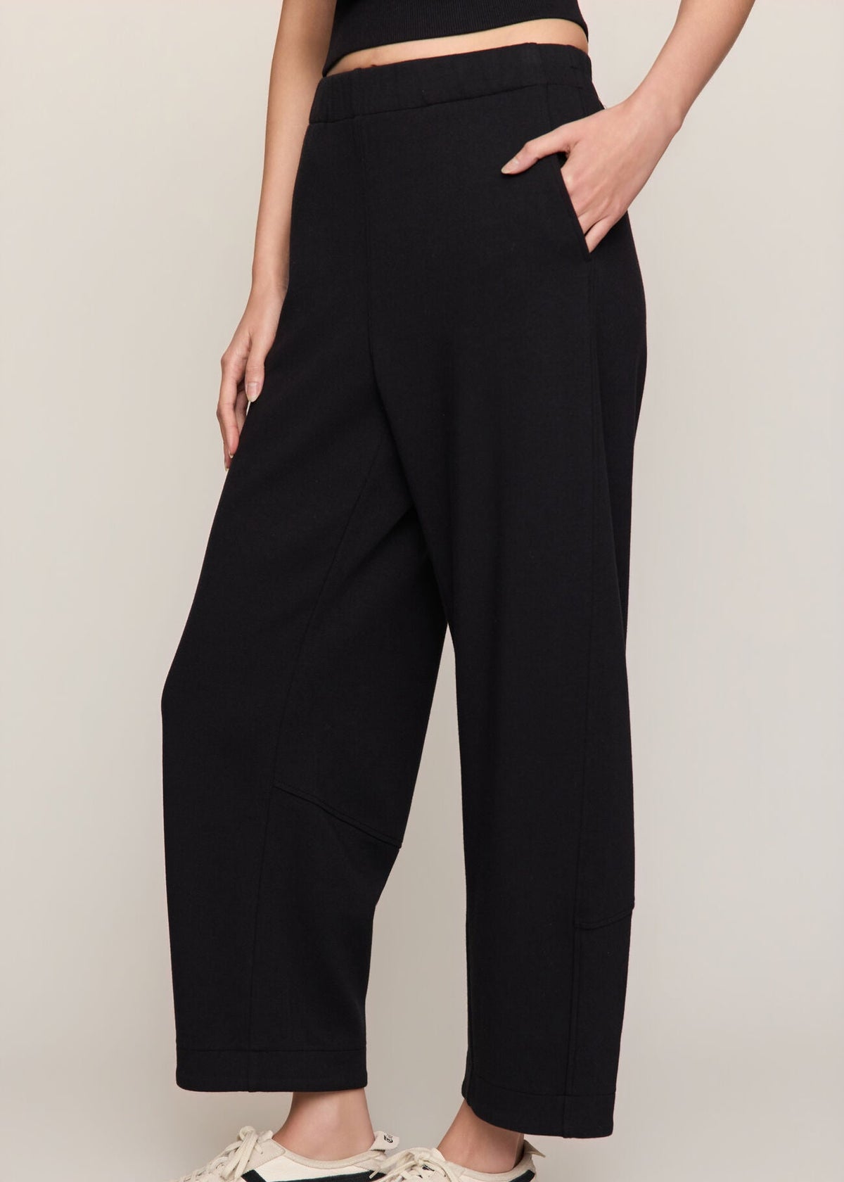A model wearing black wide-leg cropped pants with side pockets, paired with cream sneakers featuring black stripes, against a light beige studio background.