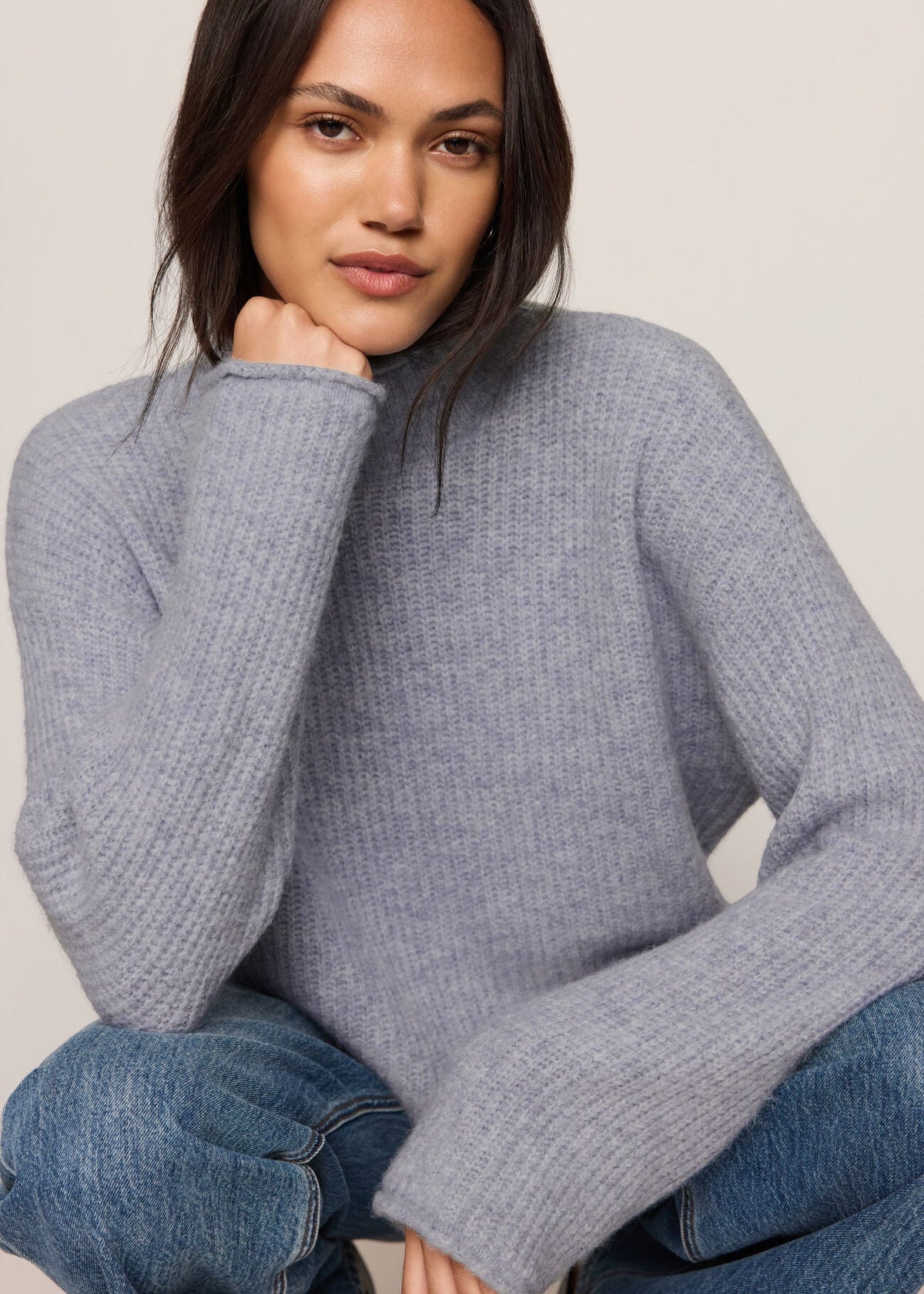 A woman wearing a light gray ribbed knit sweater and blue jeans, posing against a neutral beige background.