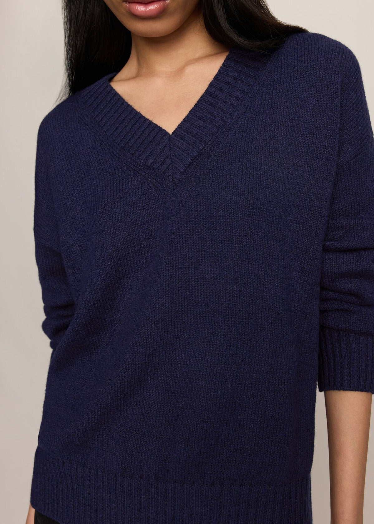 A close-up of a woman wearing a navy blue ribbed knit sweater with a V-neck and three-quarter sleeves, set against a plain light beige background.
