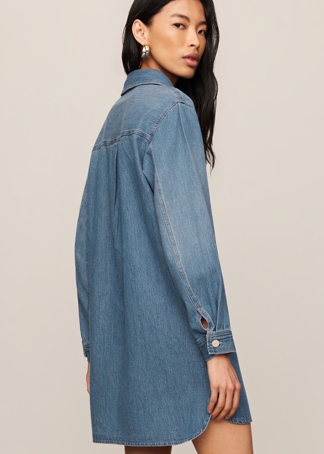 A woman modeling an oversized blue denim shirt dress, shown from a side-back angle against a plain beige studio background.
