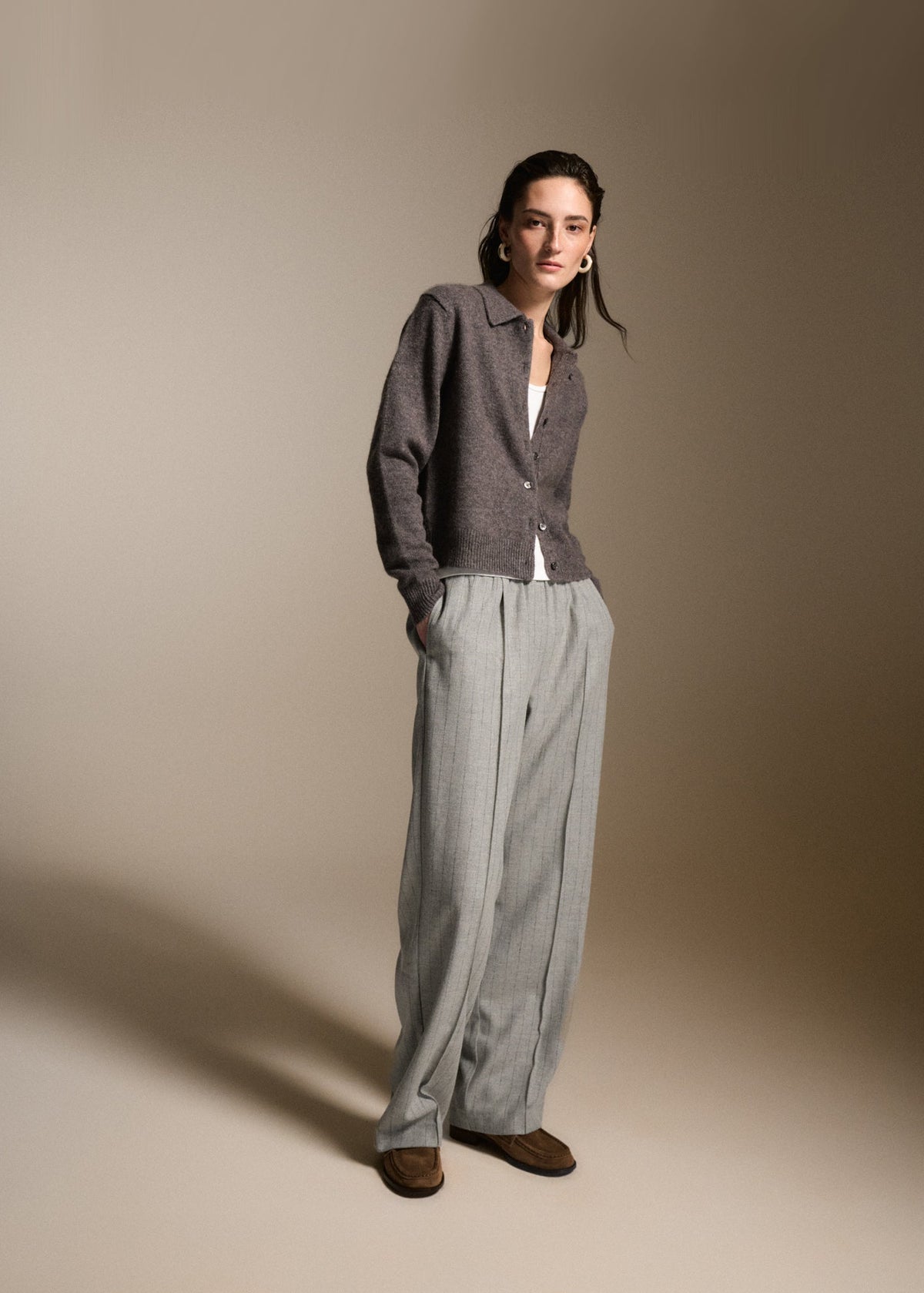 A model wearing a dark gray button-up cardigan, light gray wide-leg trousers, and brown suede loafers, standing against a warm beige studio background.