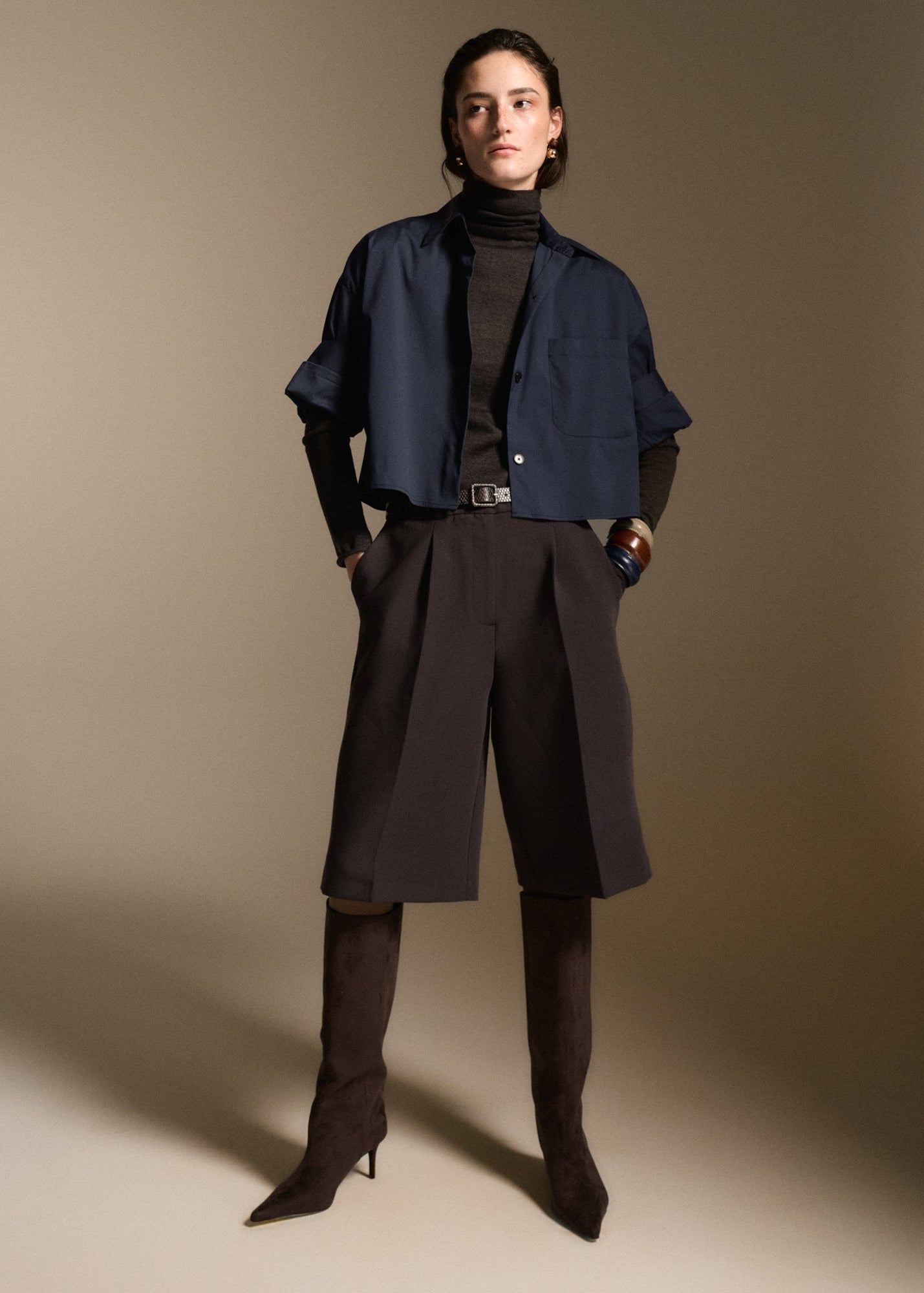 A model stands against a warm beige studio background wearing a navy cropped button-up shirt over a black turtleneck, dark pleated wide-leg shorts, and dark brown pointed-toe heeled boots.
